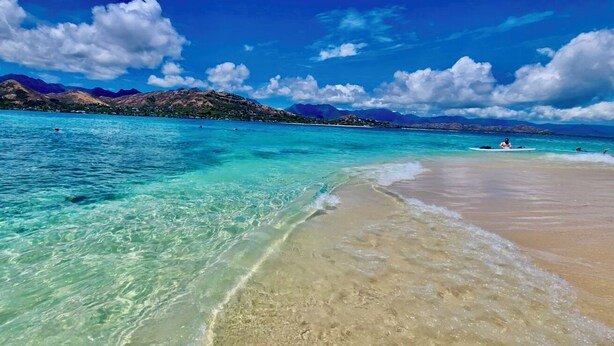 Een strand met helder turquoise water, een zandstrand en op de achtergrond bergen.