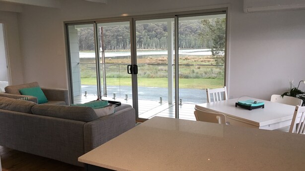 Un salon moderne avec un canapé gris, une table blanche et une vue sur un paysage naturel, visible à travers de grandes portes vitrées.