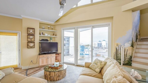 Un salón amplio con un sofá grande, un televisor y una mesa de centro de cristal. Una puerta corrediza de vidrio da a un balcón con vistas a la ciudad.