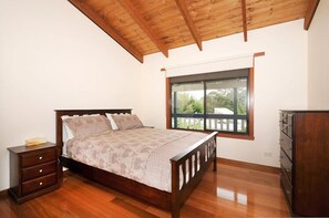 A bedroom with a wooden bed, a nightstand, a dresser, and a window with a view of greenery.