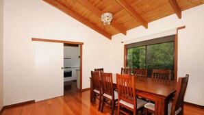 A dining area with a wooden table and chairs, a chandelier, and a view of greenery through a large window.
