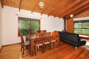 A dining area with a wooden table and chairs, a sofa, and large windows with a view of greenery.