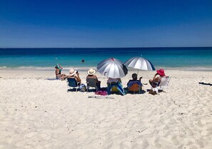 A beach scene with people sitting under umbrellas, a clear blue sky, and calm ocean waters. - Northshore apartment, separate entry, stroll to beach, quiet location. (Kallaroo)