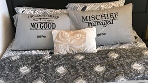 Een slaapkamer met een bed, beddengoed met Harry Potter thema, kussens met citaten uit de serie en een boekenkast met boeken en decoratieve objecten.