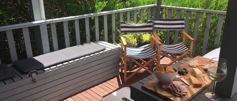 Una terraza de madera con baranda blanca, dos sillas a rayas, un banco con cojines y una mesa con comida y una copa de vino.