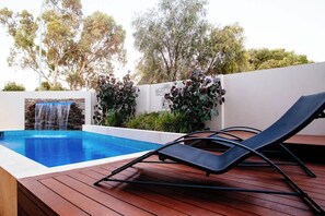 A swimming pool with a waterfall feature, a lounge chair, and a white wall with greenery.