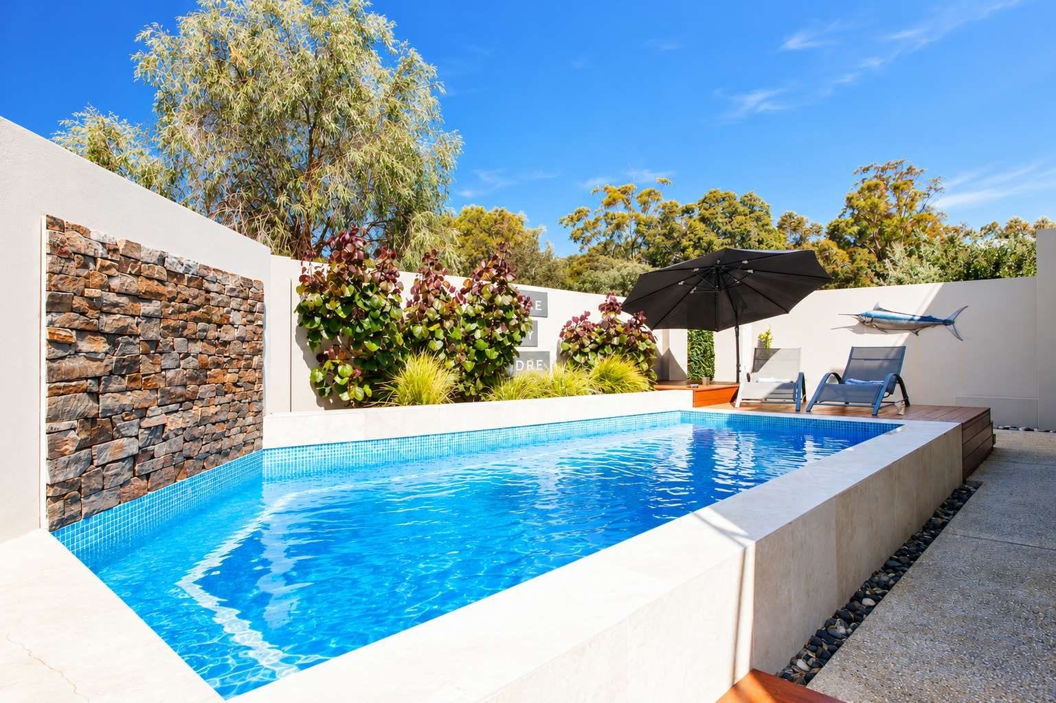 A swimming pool with a stone wall and plants, surrounded by a white fence and trees.