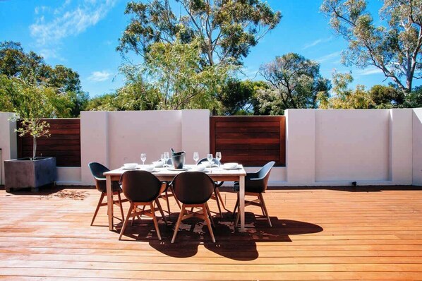 An outdoor dining area with a wooden deck, a table set for four, and a view of trees and sky.