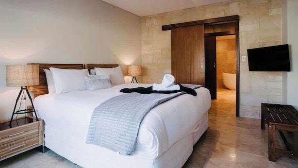 A hotel room with a large bed, bedside lamps, a flat-screen TV, a wooden bench, and a bathroom door. - Eagle Bay Beach Retreat—Steps from the Sands (Eagle Bay)