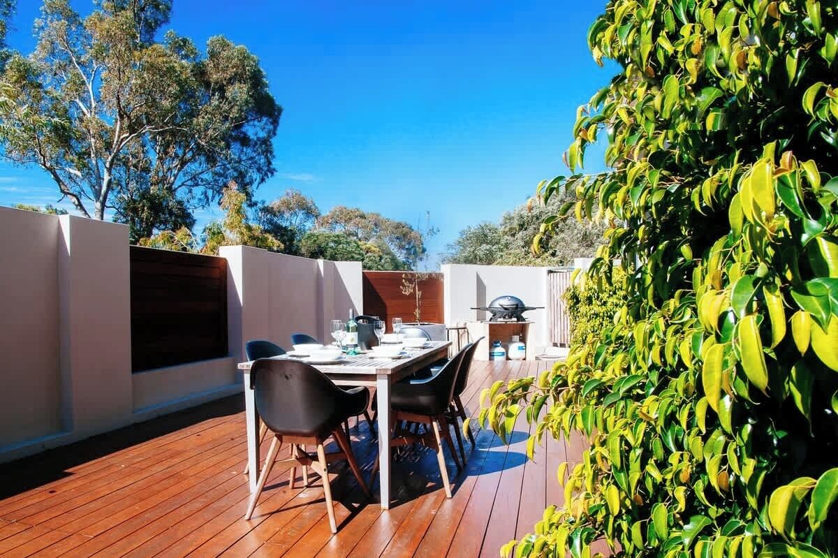 An outdoor dining area with a wooden deck, a table set for two, and a grill in the background.