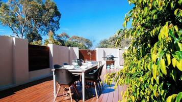 An outdoor dining area with a wooden deck, a table set for two, and a grill in the background.