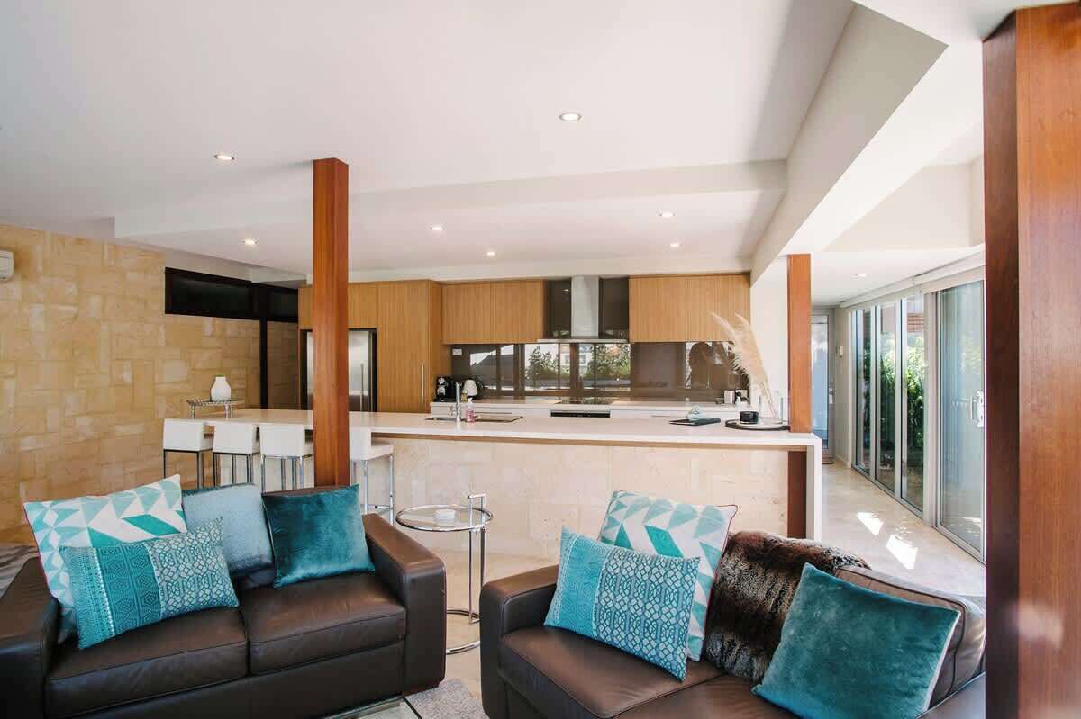 A modern kitchen with a central island, a dining area with a table and chairs, and a living area with a sofa and cushions.