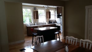 A kitchen with a central island, wooden chairs, a dining table, and a view of the outdoors through large windows. - Historic 1908 Downtown Home - Porch Swings - Mountain Views - Very Walkable (Asheville)