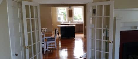 Une porte ouverte donne sur une cuisine au plancher en bois, un coin repas avec des chaises et une table, et une cheminée.