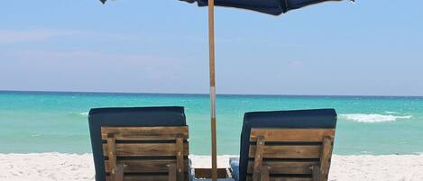 A wooden lounge chair and umbrella on a sandy beach with a clear blue sky and ocean in the background.