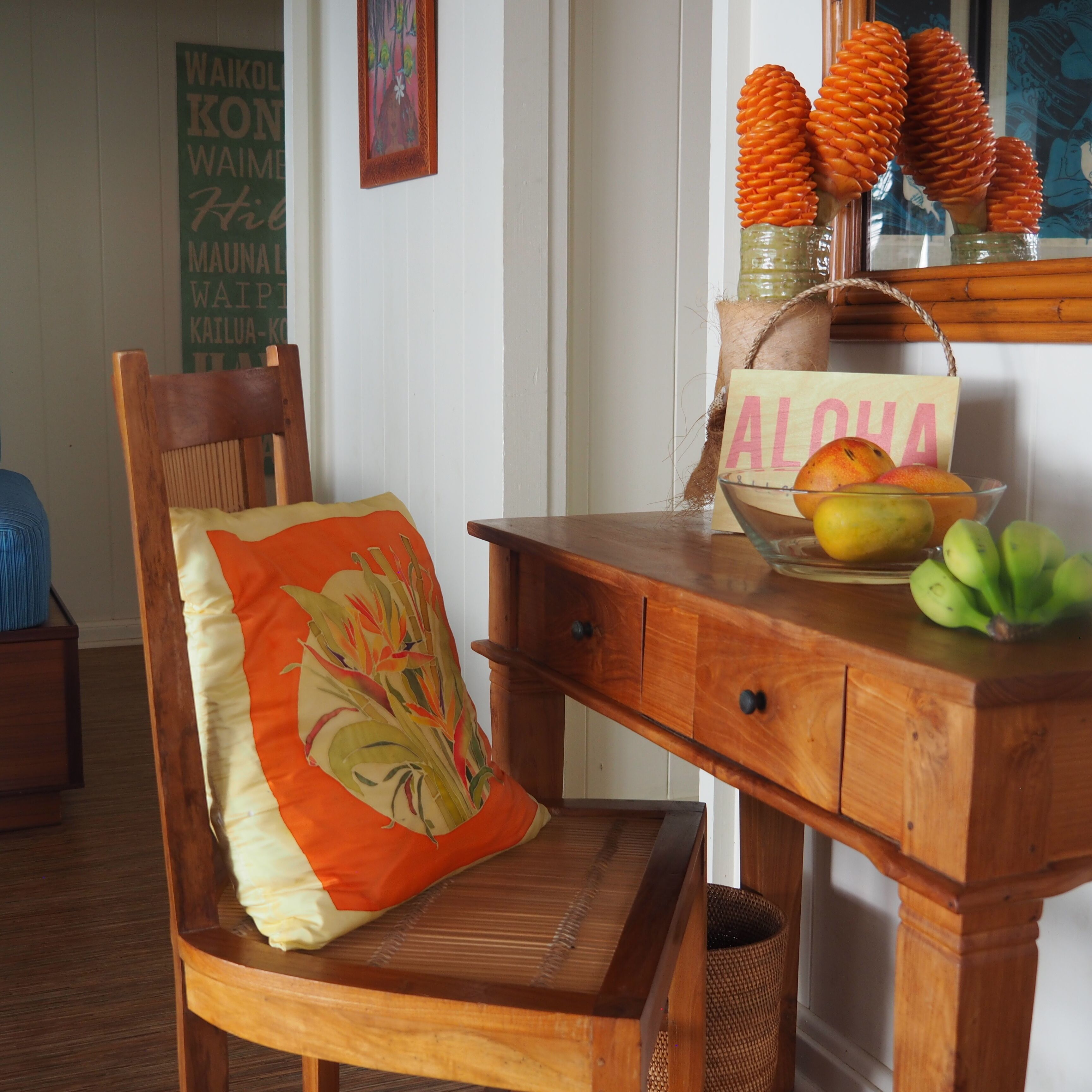 Une chaise en bois avec un coussin à motifs floraux, un bureau en bois avec un bol de fruits et un panier, et une pancarte sur laquelle est écrit le mot « ALOHA ».