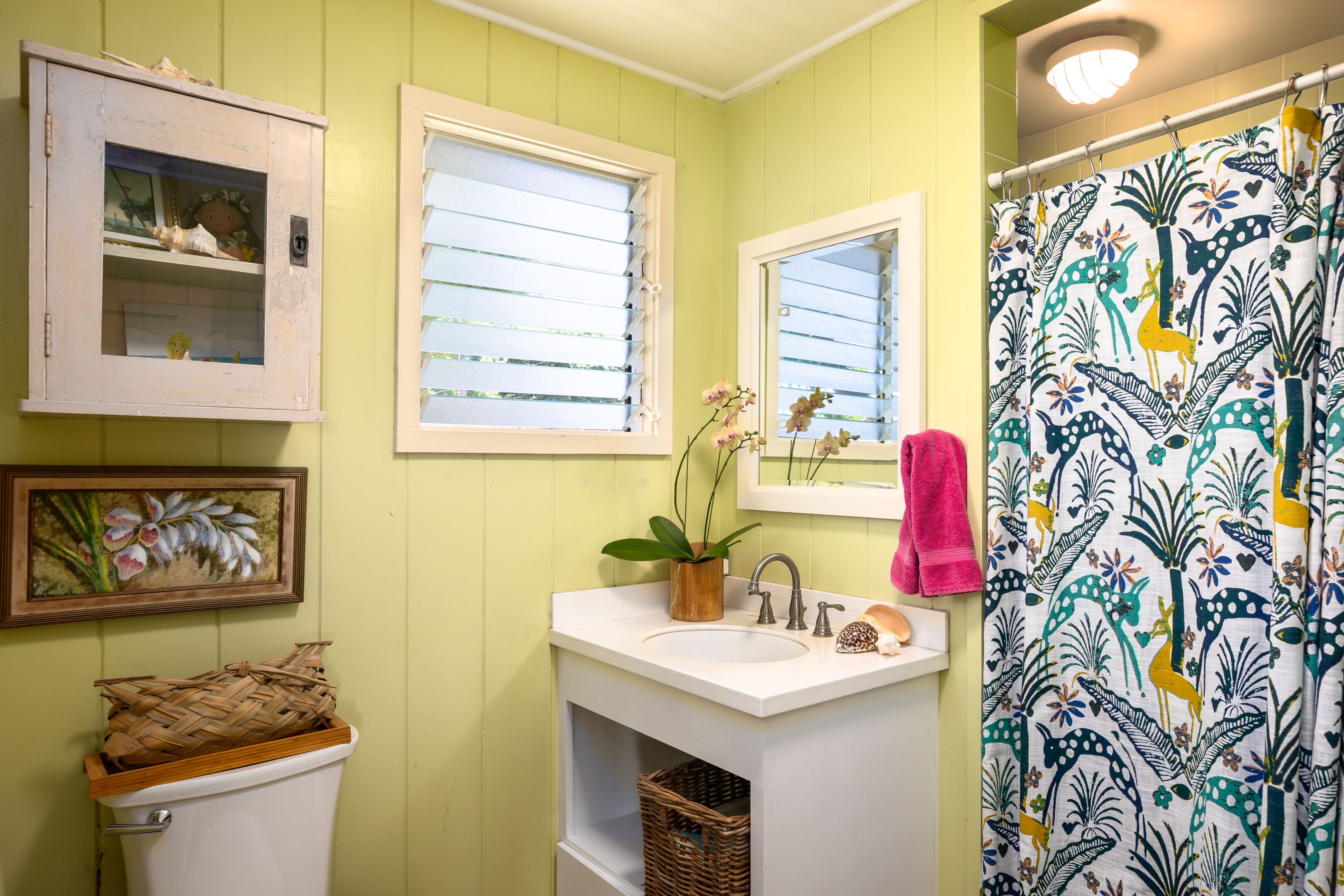 Ein Badezimmer mit einem weißen Waschbecken, einem Spiegel, einem Fenster mit Jalousien, einem Duschvorhang mit Blumenmuster und einer Toilette mit Korb darauf.