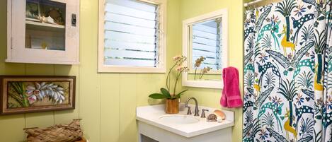Ein Badezimmer mit einem weißen Waschbecken, einem Spiegel, einem Fenster mit Jalousien, einem Duschvorhang mit Blumenmuster und einer Toilette mit Korb darauf.