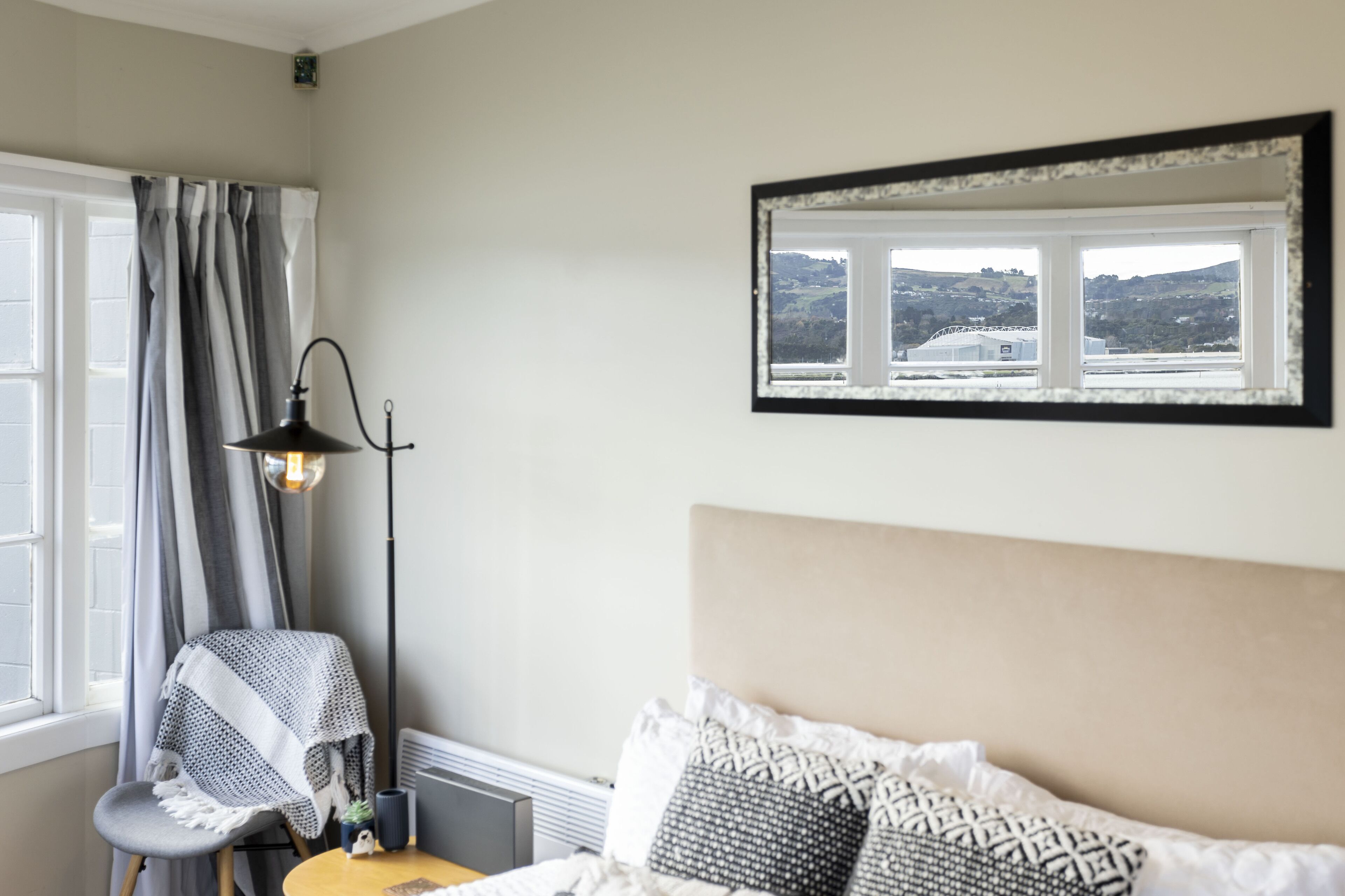 A bedroom with a bed, a chair with a blanket, a floor lamp, and a window with a view of hills.