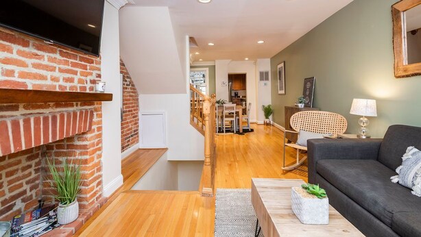 A modern living room with a brick fireplace, wooden flooring, a sofa, a dining area, and a kitchen in the background. - Amazing location for conferences and ballgames with Free Parking! (Baltimore)