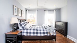 A bedroom with a checkered bedspread, a nightstand with a lamp, a dresser, and framed artwork on the wall. - Amazing location for conferences and ballgames with Free Parking! (Baltimore)