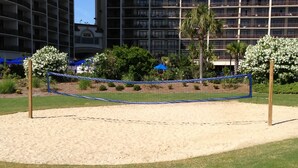Una spiaggia con un campo da pallavolo sabbioso e una rete, circondato da vegetazione e un edificio sullo sfondo.