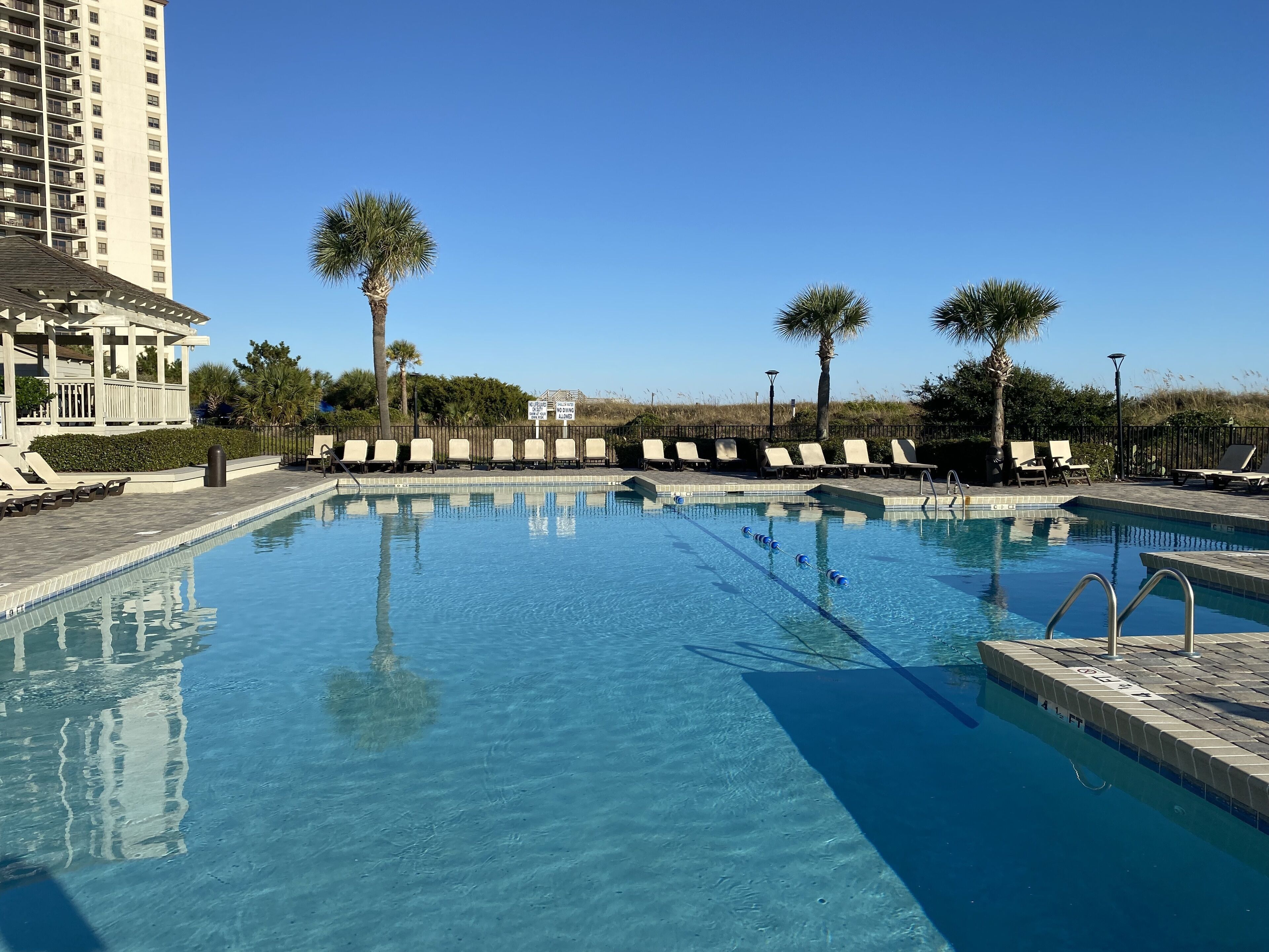 A clear swimming pool with lounge chairs and palm trees, surrounded by a fence and a building.
