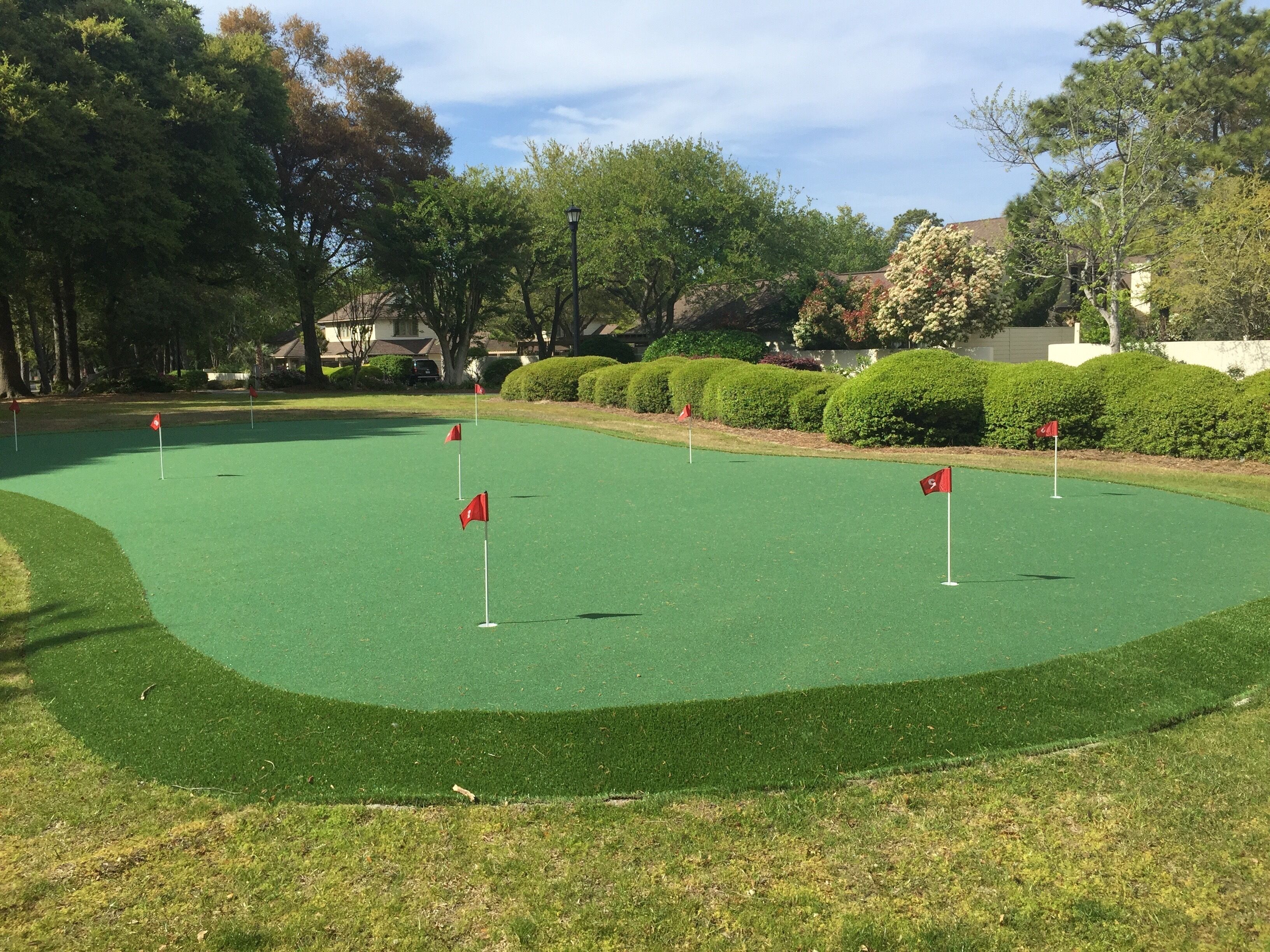 Un parcours de golf avec une pelouse artificielle verte, agrémenté de plusieurs drapeaux rouges indiquant l’emplacement des trous, et entouré d’arbres et de haies bien entretenues.