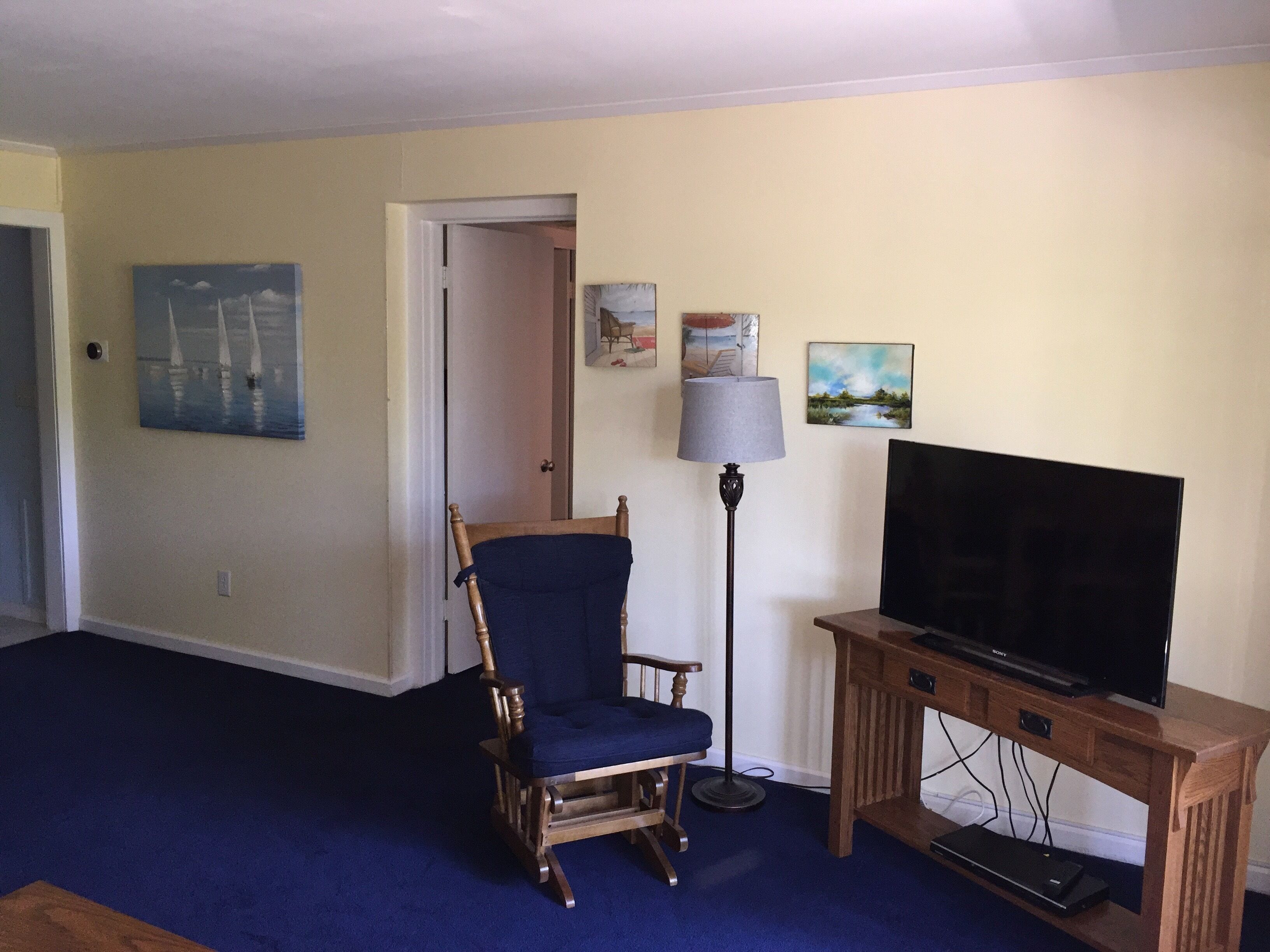 A living room with a wooden TV stand, a blue chair, a floor lamp, and artwork on the walls.