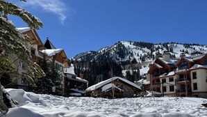Un village de montagne enneigé, avec des bâtiments traditionnels en bois, une piste de ski en arrière-plan et un ciel bleu dégagé.