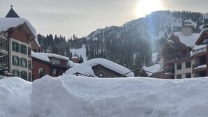 Un foyer en pierre où brûle un feu, entouré de neige, avec des bâtiments et une montagne en arrière-plan.
