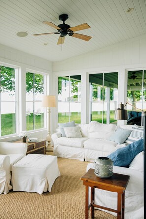 A spacious room with a white color scheme, featuring a large window, a ceiling fan, and a cozy seating area with a sofa and ottoman. - Lake Erie Lakefront Custom Built Cottage ~ Amazing Sunsets ~ Private!   (North East)