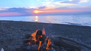 Een strand met een kampvuur op de voorgrond en een zonsondergang achterin.