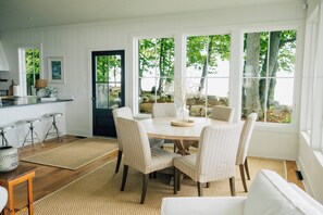 A dining area with a round table, six chairs, and a view of the outdoors through large windows. - Lake Erie Lakefront Custom Built Cottage ~ Amazing Sunsets ~ Private!   (North East)