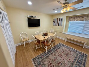 A dining area with a wooden table, white chairs, a ceiling fan, a flat-screen TV, and a window with blinds.