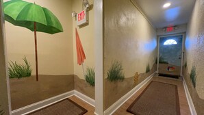 A hallway with an exit sign, a green umbrella decoration, and a door with a beach-themed window. - Beautiful Bayside Gem Accepting 3 Night Stays! (Wildwood)