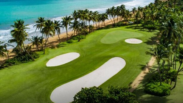 A golf course with sand bunkers, palm trees, and a beach view.