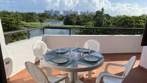 A round glass table with white plastic chairs on a terrace with a view of a lake and buildings.