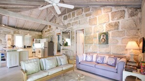 A spacious living area with a stone wall, a ceiling fan, a sofa, a coffee table, and a kitchen in the background. - Charming West Coast Cottage Near Beach (The Garden)