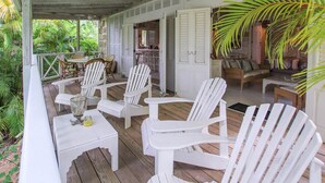 A covered porch with white Adirondack chairs, a small table with a glass, and a view of the lush greenery outside. - Charming West Coast Cottage Near Beach (The Garden)