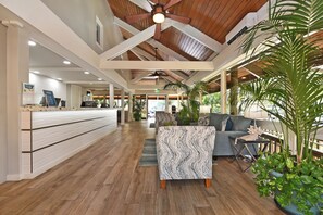 A spacious lobby with wooden flooring, a reception desk, a seating area with a patterned ottoman, and potted plants.