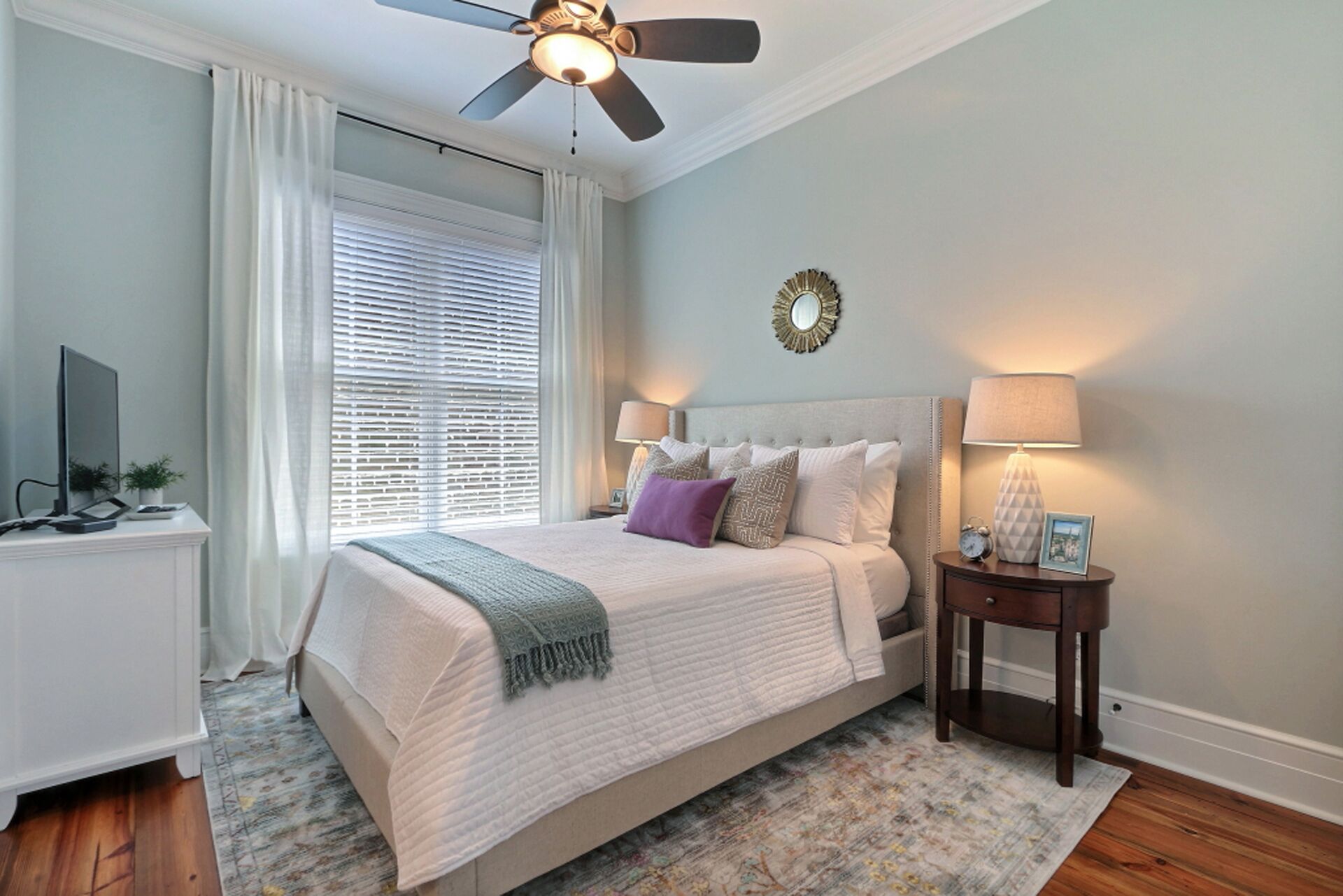 A bedroom with a bed, bedside tables, a ceiling fan, a window with blinds, a wall clock, and a decorative mirror.