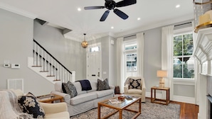 A spacious living room with a staircase, a ceiling fan, a fireplace, and a large window with blinds. - Heated Pool Access | 5*Clean | Flex Cancelation (Savannah)