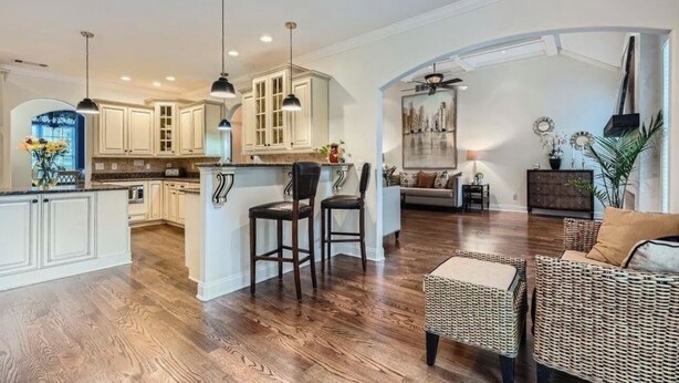 A spacious kitchen with white cabinetry and a central island, a living area with wicker furniture, and a dining area with a high ceiling and pendant lighting.
