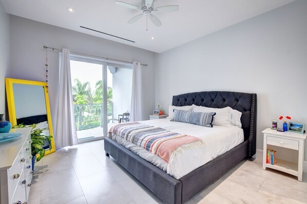 A modern bedroom with a large bed, a ceiling fan, a window with a view of palm trees, a bedside table with books, and a mirror with a yellow frame.