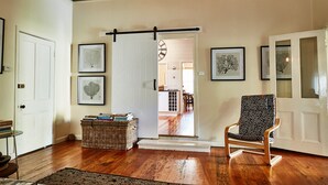 A room with hardwood flooring, a door with a sliding barn door, a chair with a patterned cushion, and framed artwork on the walls. - Inglenook Cottage Healesville (Healesville)