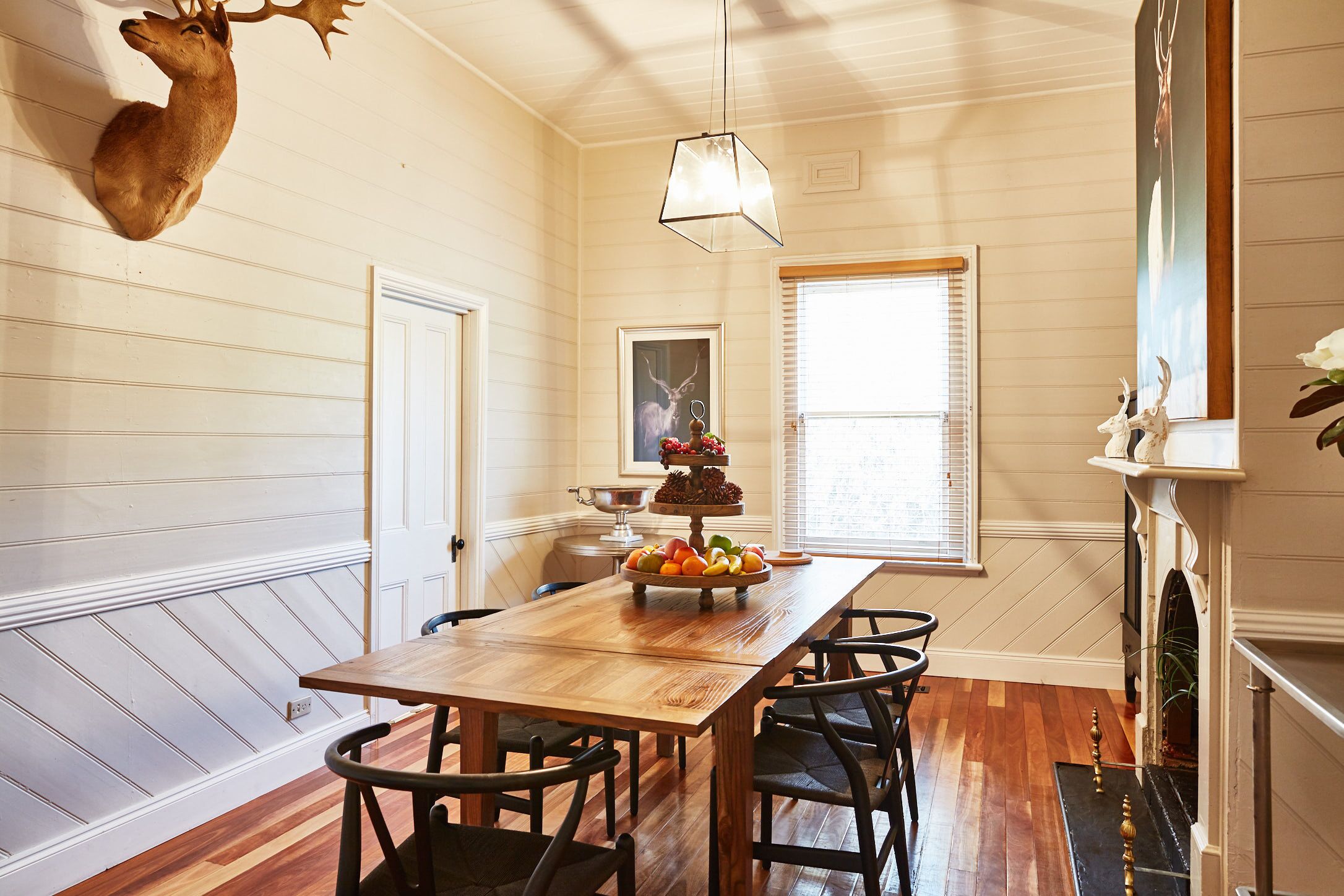 A dining room with a wooden table, black chairs, a deer head mounted on the wall, a chandelier, and a fireplace.