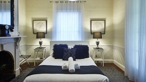 A hotel room with a large bed, two bedside lamps, a fireplace, and a window with sheer curtains. - Inglenook Cottage Healesville (Healesville)