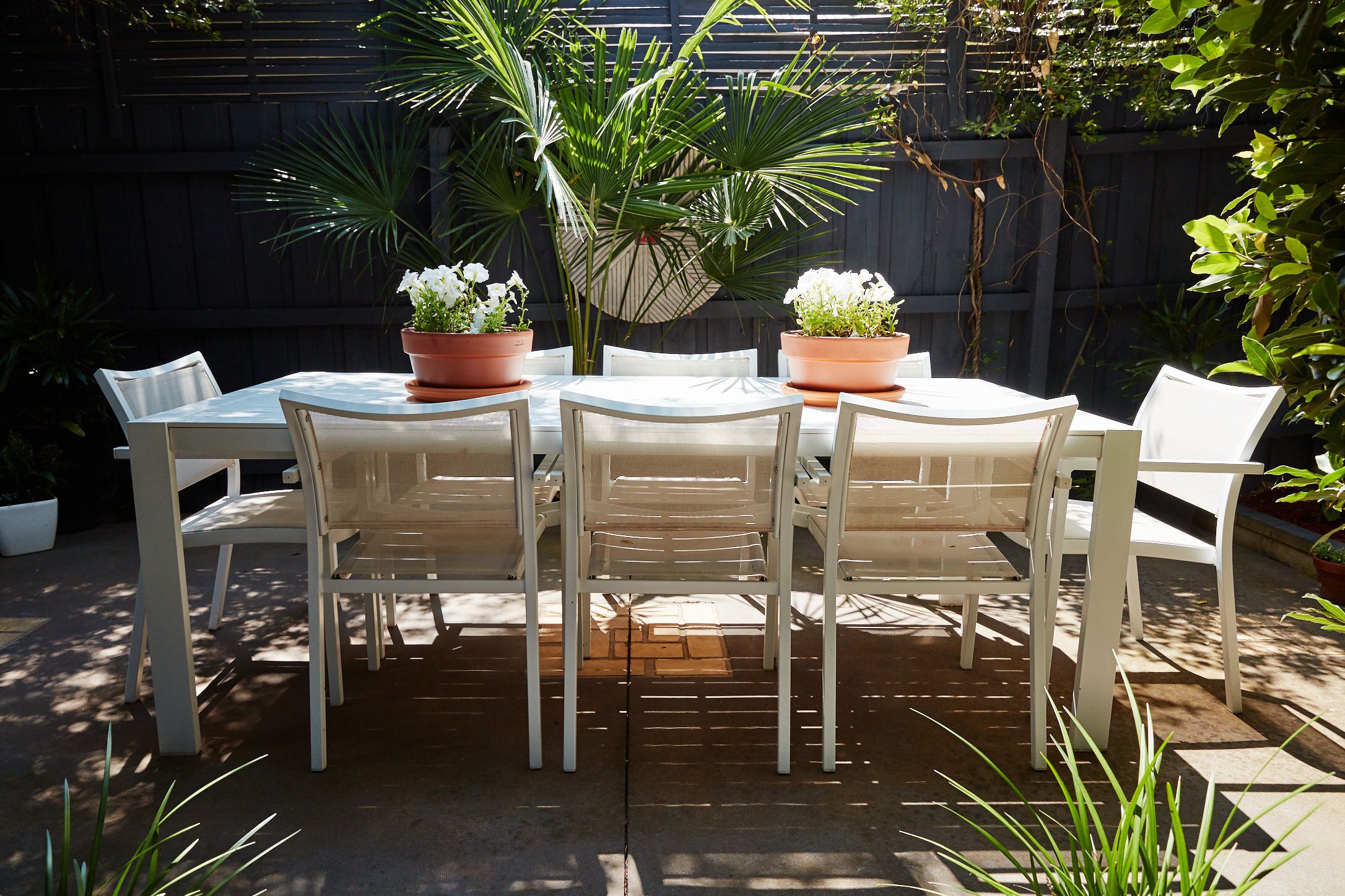 Un patio con una mesa redonda y sillas blancas, rodeado de plantas en macetas y una valla de madera.