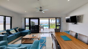 A modern living room with a sofa, coffee table, and dining area. There is a TV mounted on the wall and a view of the outdoors through large windows. - SPEEDYS SHACK   1770 - panoramic ocean & bay views (SEVENTEEN SEVENTY)